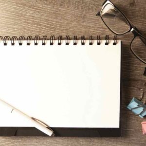 An open blank notebook with a pen, glasses and paper clips on a wooden desk