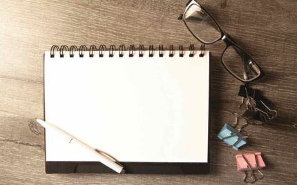 An open blank notebook with a pen, glasses and paper clips on a wooden desk