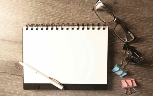 An open blank notebook with a pen, glasses and paper clips on a wooden desk