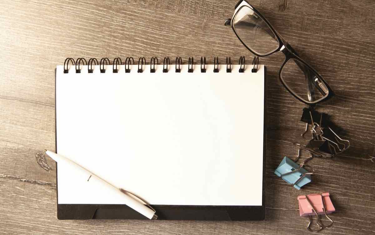 An open blank notebook with a pen, glasses and paper clips on a wooden desk