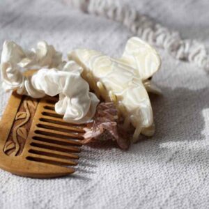 A wooden comb, a cream scrunchie and a pearl-toned claw clip resting on soft woven fabric