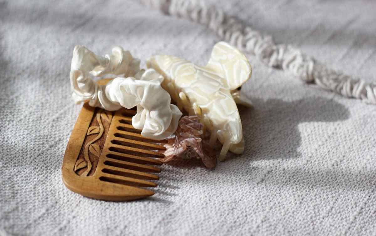 A wooden comb, a cream scrunchie and a pearl-toned claw clip resting on soft woven fabric