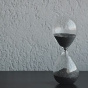 A glass hourglass with dark sand running through it, on a plain wall background