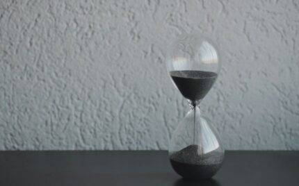 A glass hourglass with dark sand running through it, on a plain wall background