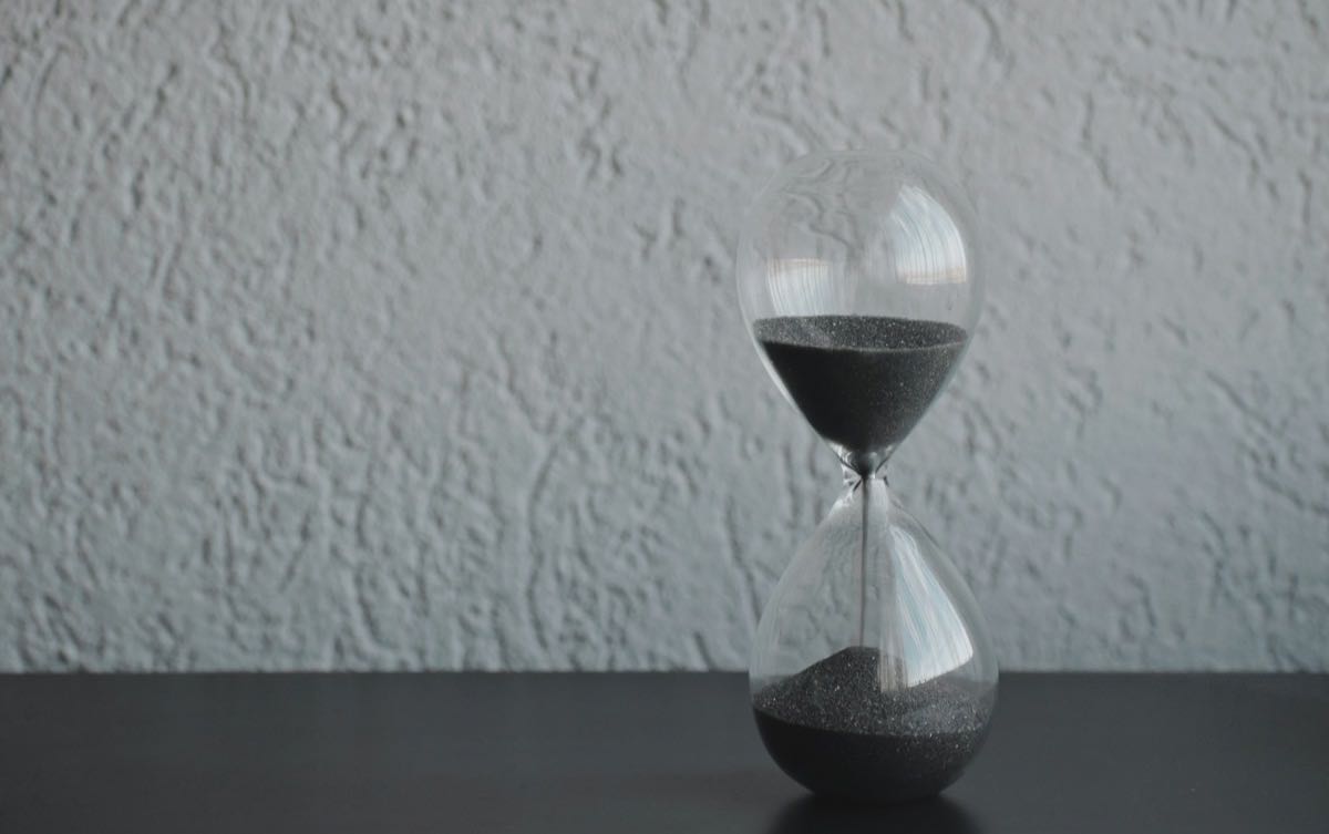 A glass hourglass with dark sand running through it, on a plain wall background